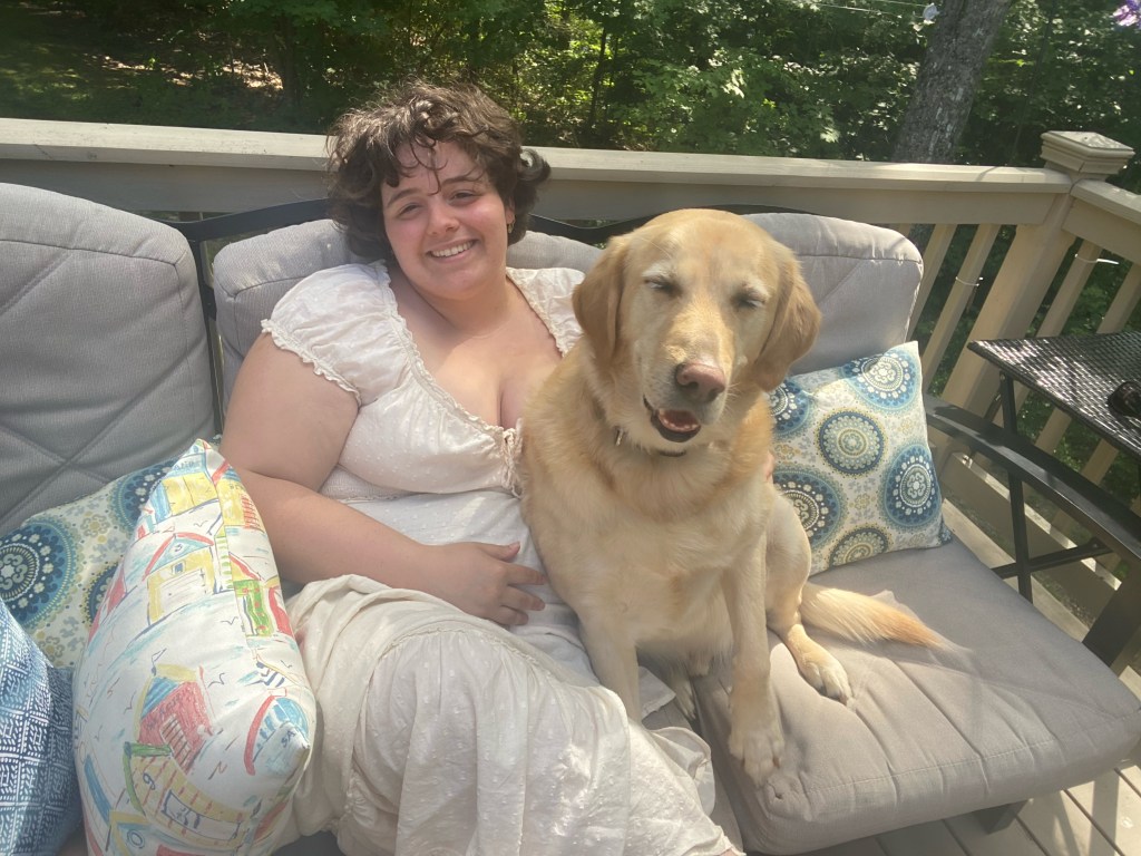 A short haired brunette woman (Alice) sits on an outdoor couch with a big yellow lab sitting in her lap dozing off. Alice wears a white dress and smiles.